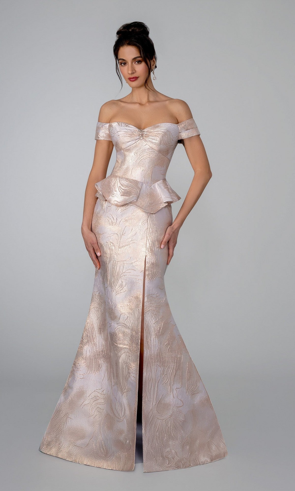 A woman wears the Stella Couture 26305 long formal dress in champagne, an off-shoulder, fitted mermaid gown with a peplum waist, front slit, and subtle floral pattern, standing against a plain background.